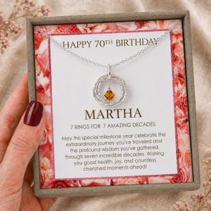 May include: A silver necklace with seven interlocking rings and a small amber-colored crystal charm, presented in a gift box with a card reading HAPPY 70TH BIRTHDAY MARTHA 7 RINGS FOR 7 AMAZING DECADES and a printed message about a milestone year.