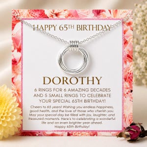 May include: A silver necklace featuring a pendant of six interlocking rings on a display card with a floral border. The text reads: HAPPY 65TH BIRTHDAY, DOROTHY, 6 RINGS FOR 6 AMAZING DECADES AND 5 SMALL RINGS TO CELEBRATE YOUR SPECIAL 65TH BIRTHDAY!