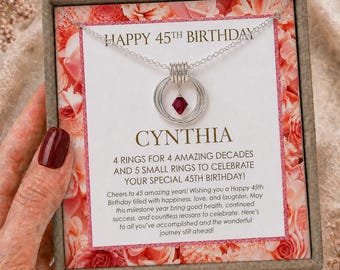 May include: A silver necklace with a pendant featuring four interlocking rings and a small red crystal bead, presented in a box with a card that reads HAPPY 45TH BIRTHDAY CYNTHIA, 4 RINGS FOR 4 AMAZING DECADES AND 5 SMALL RINGS TO CELEBRATE YOUR SPECIAL 45TH BIRTHDAY!