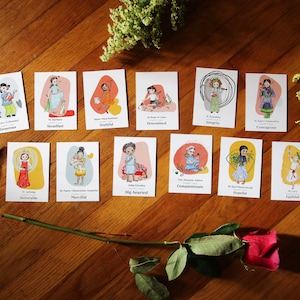 Puede incluir: Un conjunto de 14 tarjetas ilustradas con diferentes figuras femeninas con una palabra que describe su rasgo de carácter. Las tarjetas están sobre una superficie de madera con una rosa y follaje verde.