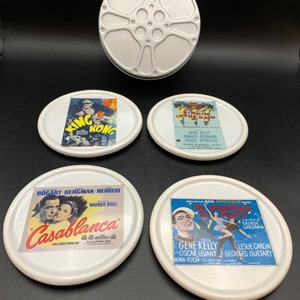 May include: A set of four white ceramic coasters featuring classic movie posters, including "King Kong," "Singin' in the Rain," "Casablanca," and "An American in Paris." A silver film reel container is above the coasters.
