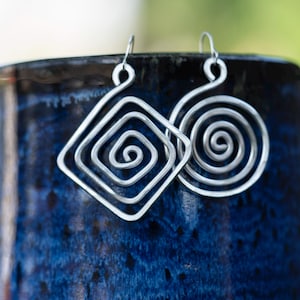 May include: Silver-toned earrings with a geometric design. One earring is a square spiral, the other a circular spiral. The earrings are displayed against a dark blue, textured background, with a blurred, natural backdrop.
