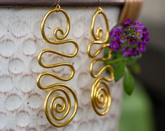 May include: A pair of gold-toned wire earrings featuring a continuous, flowing design that incorporates multiple loops and a prominent circular spiral at the base. The metal wire is shaped into an elongated, sinuous pattern with a smooth, polished finish.