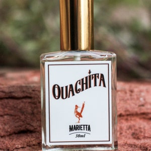 May include: A clear glass bottle with a gold cap and a white label. The label has the text "Ouachita" in black, a red bird illustration, and the text "Marietta 30ml".