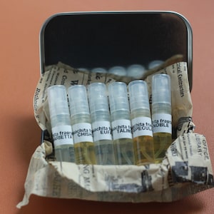 May include: A tin box filled with six small clear glass bottles containing perfume samples. The bottles are labeled with the names "fragrancita", "talisma", "speculum", "noble", "chish", and "marietta".