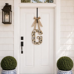 May include: A white front door with a black handle and a decorative wreath in the shape of the letter "G" made of oyster shells. Two blue and white patterned planters with green bushes flank the door. A black lantern hangs on the wall.