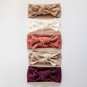 May include: Five different coloured knotted headbands. The headbands are made of a soft, textured fabric. The colours are brown, white, pink, beige, and burgundy.