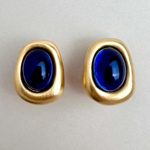 May include: A pair of earrings featuring a smooth, matte gold-toned metal frame with an irregular, rounded shape. Each earring is centred with a large, polished, deep blue oval cabochon stone that reflects light on its curved, glossy surface.