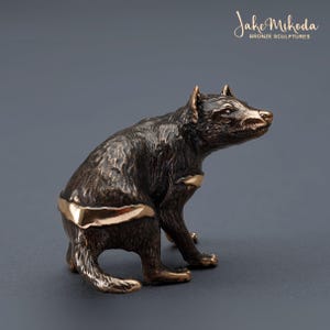 Puede incluir: Escultura de bronce de un demonio de Tasmania sentado en sus ancas. La escultura está detallada con un acabado texturizado y una franja dorada alrededor del cuerpo del animal. La escultura está firmada "Jake Mikoda Bronze Sculptures".