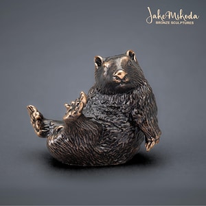 May include: A bronze sculpture of a bear sitting on its haunches with its paws outstretched. The bear has a playful expression on its face and appears to be relaxed. The sculpture is made of a dark bronze material and has a smooth, polished finish.