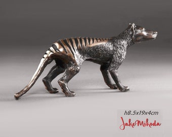 May include: Bronze sculpture of a Tasmanian tiger, also known as a thylacine, standing on a gray surface. The sculpture is 8.5 inches tall, 19 inches long, and 4 inches wide. The sculpture is signed "Jake Mikoda".