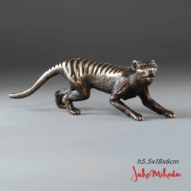 May include: Bronze sculpture of a Tasmanian tiger, also known as a thylacine, in a crouching pose. The sculpture is 5.5 inches tall, 18 inches long, and 6 inches wide. The sculpture is signed "Jake Mikoda".