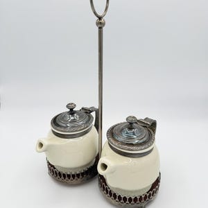 May include: A vintage cream-colored ceramic salt and pepper set with silver-toned lids and trim. The set includes two small, lidded pots with spouts, each resting in a decorative metal holder. A tall, slender handle connects the two pots.