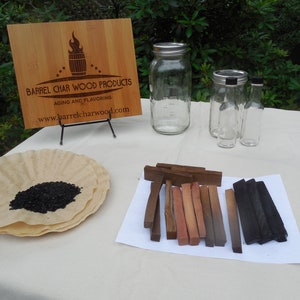 May include: A wooden sign with the text "Barrel Char Wood Products Aging and Flavoring www.barrelcharwood.com" with a small barrel and flame graphic.  A white table with a variety of glass jars and bottles, a plate with black wood chips, and a selection of wood sticks in various shades of brown and black.