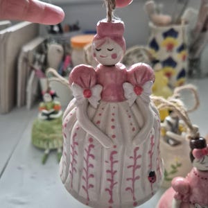 May include: A ceramic bell-shaped ornament with a pink and white dress design, featuring a ladybug detail. The ornament is suspended by a natural twine cord. The figure has a pink top and a smiling face. Other similar ornaments are visible in the background.