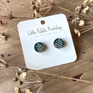 Puede incluir: Pendientes de botón redondos con fondo verde oscuro y diseño de helecho blanco. Los pendientes se muestran en una tarjeta blanca con el texto "Little Pebble Printshop". La tarjeta está sobre una superficie de madera.