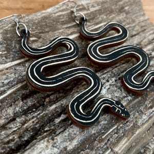 May include: A pair of black and white snake earrings made of wood. The earrings are shaped like a snake with a white stripe down the middle. The earrings are hanging from silver hooks.