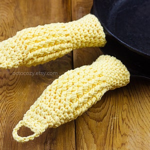May include: Two yellow crocheted pot holders with a black cast iron skillet in the background. The pot holders are made with a simple stitch pattern and have a loop at the top for hanging.
