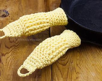 May include: Two yellow crocheted pot holders with a black cast iron skillet in the background. The pot holders are made with a simple stitch pattern and have a loop at the top for hanging.