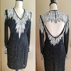 May include: A black, long-sleeved dress with a V-neck and a low back. The dress is embellished with white and silver beading in a feather-like pattern on the bodice and sleeves. The dress is knee-length and has a fitted silhouette.