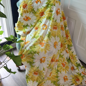 Puede incluir: Una tela blanca con un gran estampado floral de girasoles amarillos y naranjas con hojas verdes. La tela está drapeada sobre una silla azul claro. El fondo incluye una puerta blanca, una ventana y una pared azul oscuro.