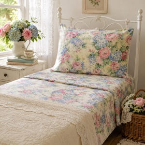 May include: A bed featuring a cream-colored sheet and pillowcase set adorned with a floral pattern of pink roses and blue hydrangeas. The bedding includes a textured cream-colored throw blanket with a decorative lace trim along the edge.