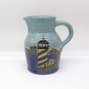 May include: A ceramic pitcher with a blue and white lighthouse design. The pitcher has a handle and a spout. The lighthouse is painted with black and yellow stripes, and the scene includes a small house and water.