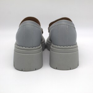 Pu&ograve; includere: Un paio di scarpe grigie con plateau e suola spessa. Le scarpe hanno la punta arrotondata e un cinturino sulla parte superiore. La suola platform &egrave; alta circa 7.6 cm. Le scarpe sono realizzate in materiale sintetico liscio.