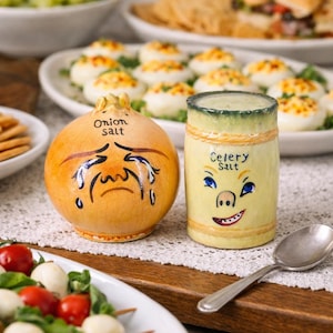 Może przedstawiać: Kapryśny zestaw solniczki i pieprzniczki ceramicznej. Solniczka do cebuli ma kształt cebuli z płaczącą twarzą. Pieprzniczka do selera to żółty walec z uśmiechniętą twarzą. Zestaw stoi na stole z jedzeniem.