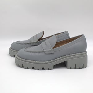 Pu&ograve; includere: Un paio di mocassini grigi con plateau. Queste scarpe presentano una suola spessa e carrarmato e un classico design a mocassino. I tacchi misurano circa 5 cm.