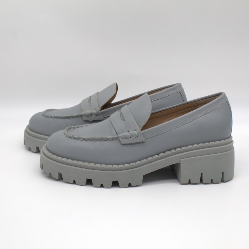 Pu&ograve; includere: Un paio di mocassini grigi con plateau. Queste scarpe presentano una suola spessa e carrarmato e un classico design a mocassino. I tacchi misurano circa 5 cm.