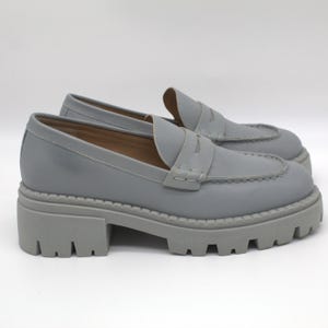 Pu&ograve; includere: Un paio di mocassini grigi in pelle con suola carrarmato. Le scarpe hanno un classico design penny loafer con tomaia cucita e punta arrotondata. Le suole sono alte circa 5 cm.