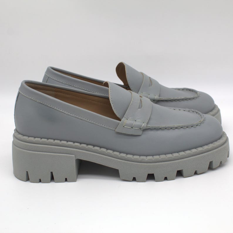 Pu&ograve; includere: Un paio di mocassini grigi in pelle con suola carrarmato. Le scarpe hanno un classico design penny loafer con tomaia cucita e punta arrotondata. Le suole sono alte circa 5 cm.