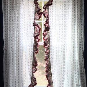 May include: A window is framed by white lace curtains with a floral pattern. Ruffled valances with a floral print and burgundy trim hang on either side of the window. The window looks out onto a green landscape.