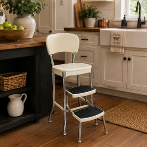 May include: A vintage-style step stool chair featuring a cream-colored seat and backrest with a chrome metal frame. The stool includes two retractable steps with black ribbed treads, providing a functional seating and climbing solution.