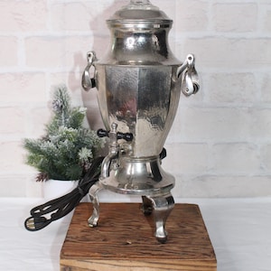 May include: A vintage silver-toned electric samovar with a clear glass lid. The samovar has a hammered texture, black handles, and a spigot. It stands on ornate legs and is placed on a wooden box. A black power cord is visible.
