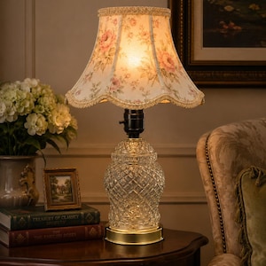 May include: A table lamp with a clear, diamond-patterned glass base and a gold-toned metal circular stand. The lamp features a bell-shaped fabric lampshade decorated with a pink rose floral pattern and finished with a scalloped gold trim edge.