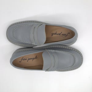 Pu&ograve; includere: Un paio di mocassini in pelle grigio chiaro con design a cinturino penny. Le scarpe hanno una punta arrotondata e una suola spessa e strutturata. Il marchio "free people" &egrave; stampato sulla soletta di ogni scarpa.