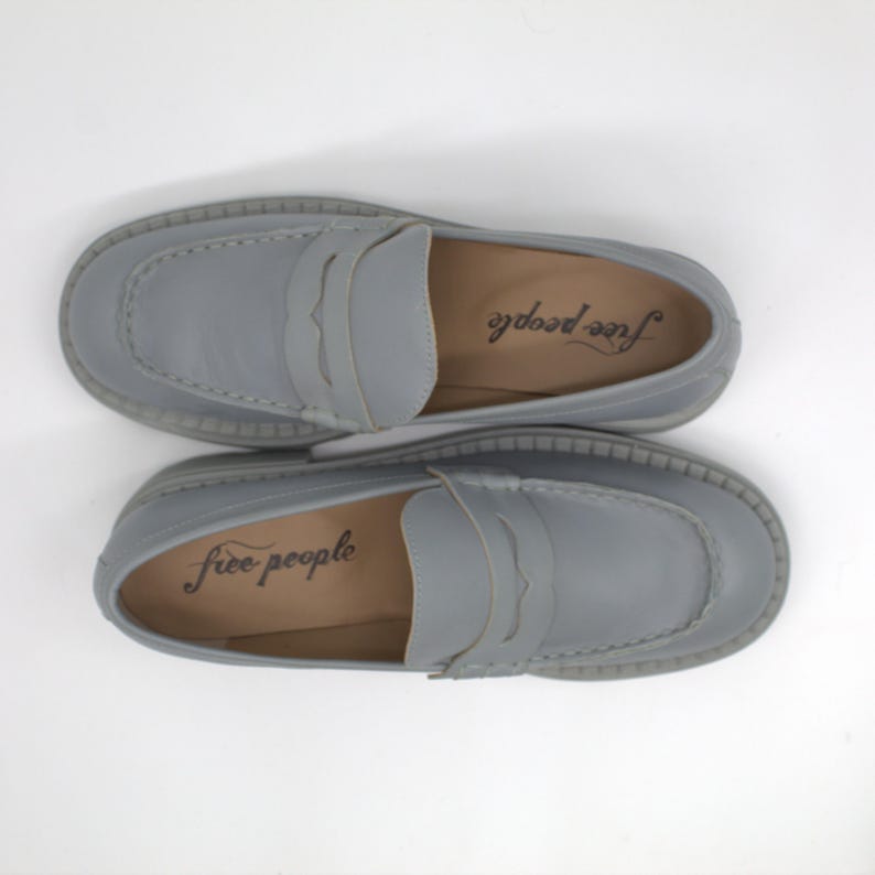 Pu&ograve; includere: Un paio di mocassini in pelle grigio chiaro con design a cinturino penny. Le scarpe hanno una punta arrotondata e una suola spessa e strutturata. Il marchio "free people" &egrave; stampato sulla soletta di ogni scarpa.