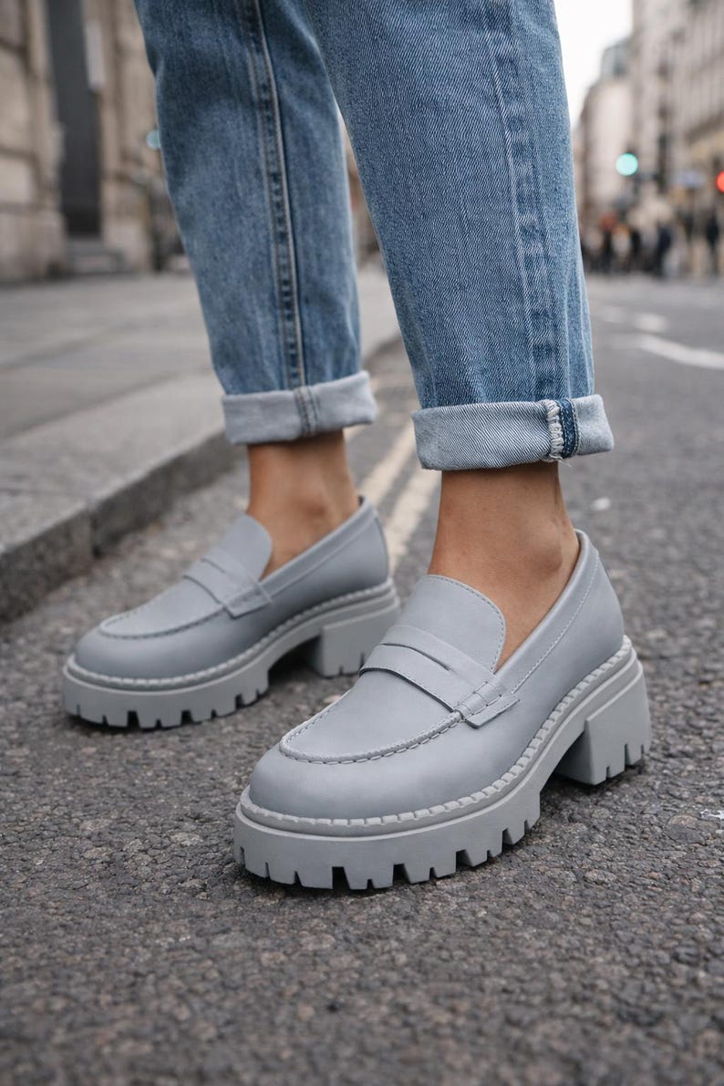 Pu&ograve; includere: Mocassini in pelle grigia con suola spessa e carrarmato. Le scarpe hanno un classico design penny loafer con punta arrotondata e cinturino sul collo del piede. Le scarpe sono abbinate a jeans blu.