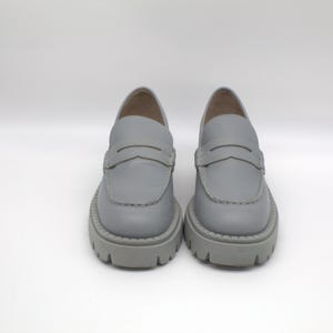 Pu&ograve; includere: Un paio di mocassini in pelle grigio chiaro con suola spessa e carrarmato. Le scarpe presentano un classico design penny loafer con una fascia sul collo del piede. Le suole spesse e grigie offrono un look moderno ed elegante.