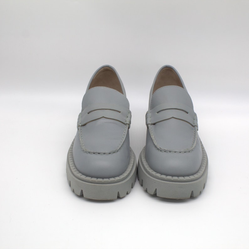 Pu&ograve; includere: Un paio di mocassini in pelle grigio chiaro con suola spessa e carrarmato. Le scarpe presentano un classico design penny loafer con una fascia sul collo del piede. Le suole spesse e grigie offrono un look moderno ed elegante.