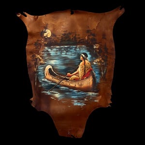 May include: Hand-painted artwork on tanned hide depicting a person in a canoe on a lake under a full moon. The artwork features a person in a canoe with a paddle, surrounded by trees and water. The hide is brown with a unique shape.