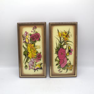 May include: Two framed floral art pieces. Each features a bouquet of yellow, pink, and white flowers with green leaves and a butterfly against a light yellow background. The frames are a warm brown color.