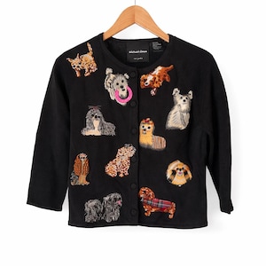 May include: A black long-sleeve cardigan featuring a variety of embroidered dog designs across the front. The garment has a round neckline, a button-down front closure, and a label at the neck that reads Michael Simon. The dogs are depicted in various colors and poses, including one holding a pink ring.