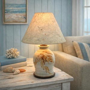 May include: A ceramic table lamp with a beige lampshade and a cream-colored base decorated with brown leaf designs. The lamp sits on a wooden table, creating a warm, inviting ambiance. The lamp's height is approximately 18 inches.