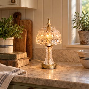 May include: A table lamp featuring a clear, faceted glass shade and a matching glass base with a metallic gold-toned finish. The lamp emits a warm, glowing light from within, highlighting the intricate geometric patterns of the glass construction.