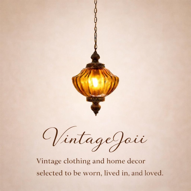 Pu&ograve; includere: Una lampada a sospensione in stile vintage con un paralume in vetro ambrato caldo e dettagli in metallo ornati. La lampada &egrave; sospesa a una catena ed emette una luce soffusa e invitante. Il testo "Vintage Joii" e una descrizione del negozio sono sotto.