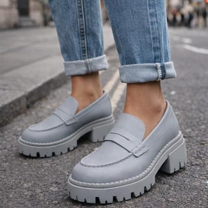 Pu&ograve; includere: Mocassini platform grigio chiaro con suola carrarmato. Le scarpe hanno un classico design penny loafer con punta arrotondata e tacco spesso. Le scarpe sono abbinate a jeans blu.