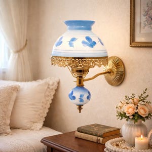 Puede incluir: Una lámpara de pared de estilo vintage con una pantalla de cristal blanca y azul con estampado floral. La lámpara tiene una base y un brazo de metal color latón y está montada en una pared. La lámpara está cerca de una mesita de noche con un jarrón de flores y una vela encendida.